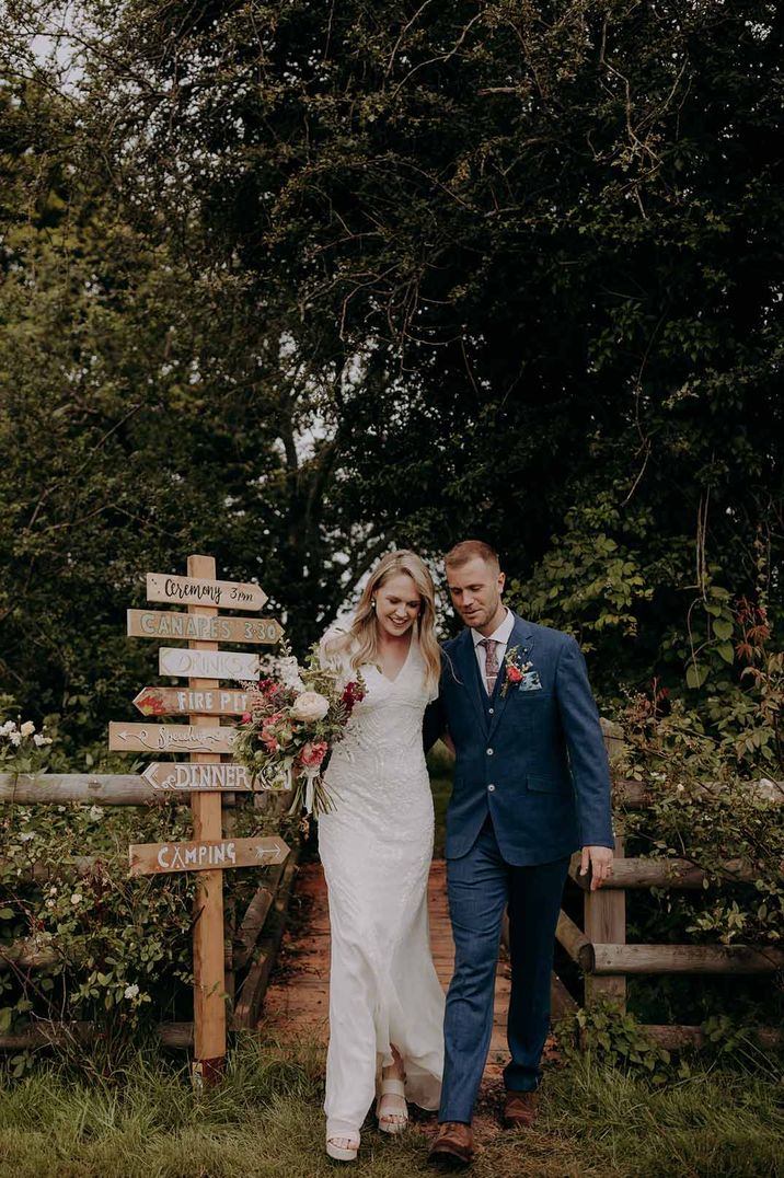 Bride in short sleeved embellished wedding dress and groom in dark suit standing by DIY wedding sign