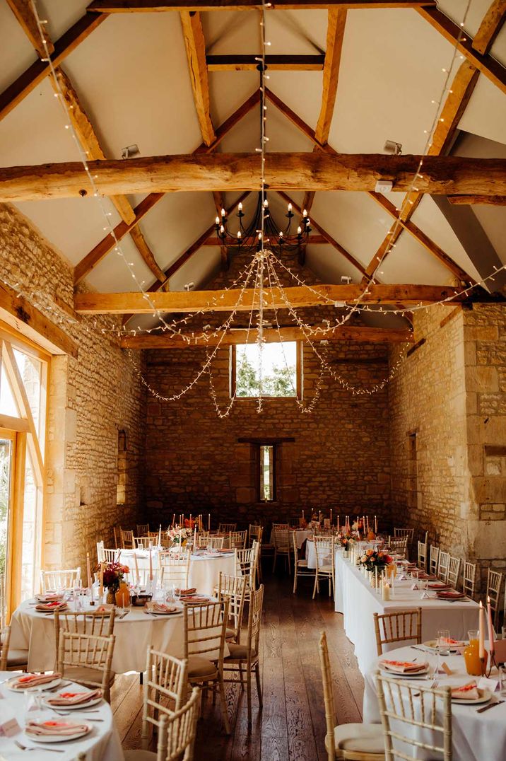 Wedding reception room at The Barn at Upcote with neutral white wedding tablescapes, exposed brick and fairy lights hung on the ceiling beams