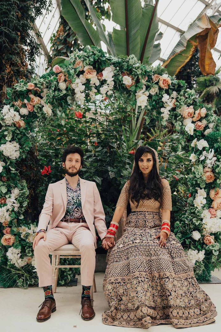 Bride in traditional Hindu wedding garments and groom in pale suit and colourful shirt standing by large exotic plants at green house wedding venues Sefton Park Palm House 