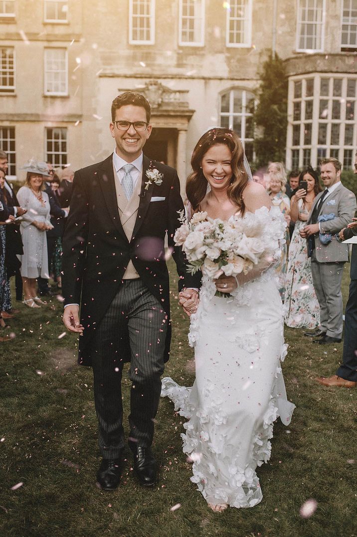 Groom in a morning suit with cream waistcoat and blue tie with the bride in an off the shoulder floral wedding dress having a confetti exit 