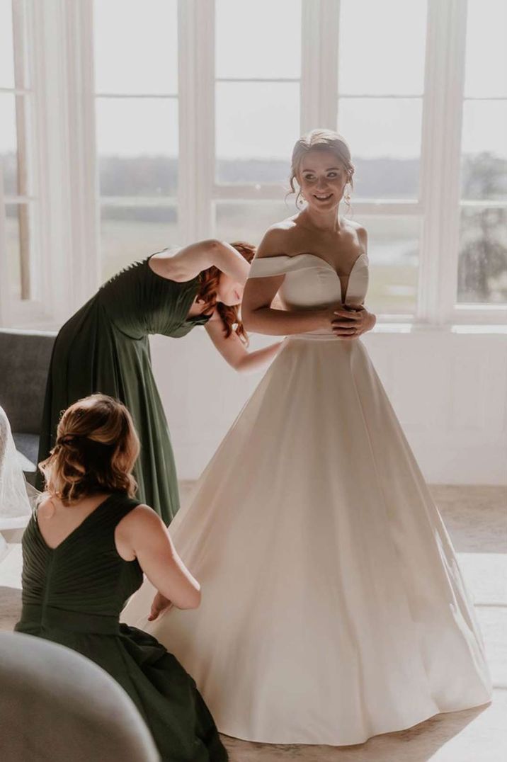 Bride in deep v neck off the shoulder princess wedding dress getting help from her friends in black bridesmaid dresses 