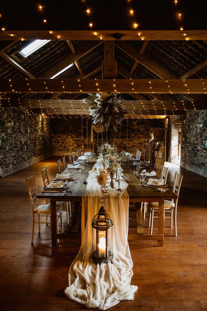 Wooden tables covered in white table runner and warm candlelight with pampas grass installation and dried wedding flowers 