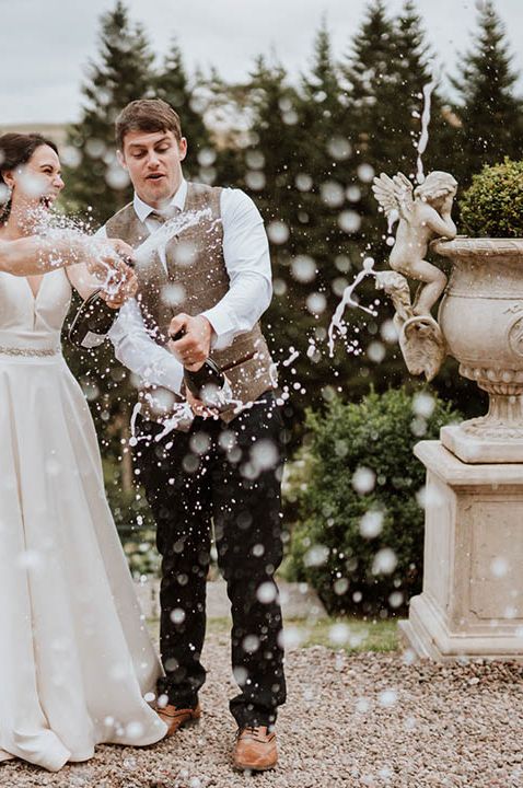 Bride a princess wedding dress and groom in a wool waistcoat popping champagne at their country house wedding 