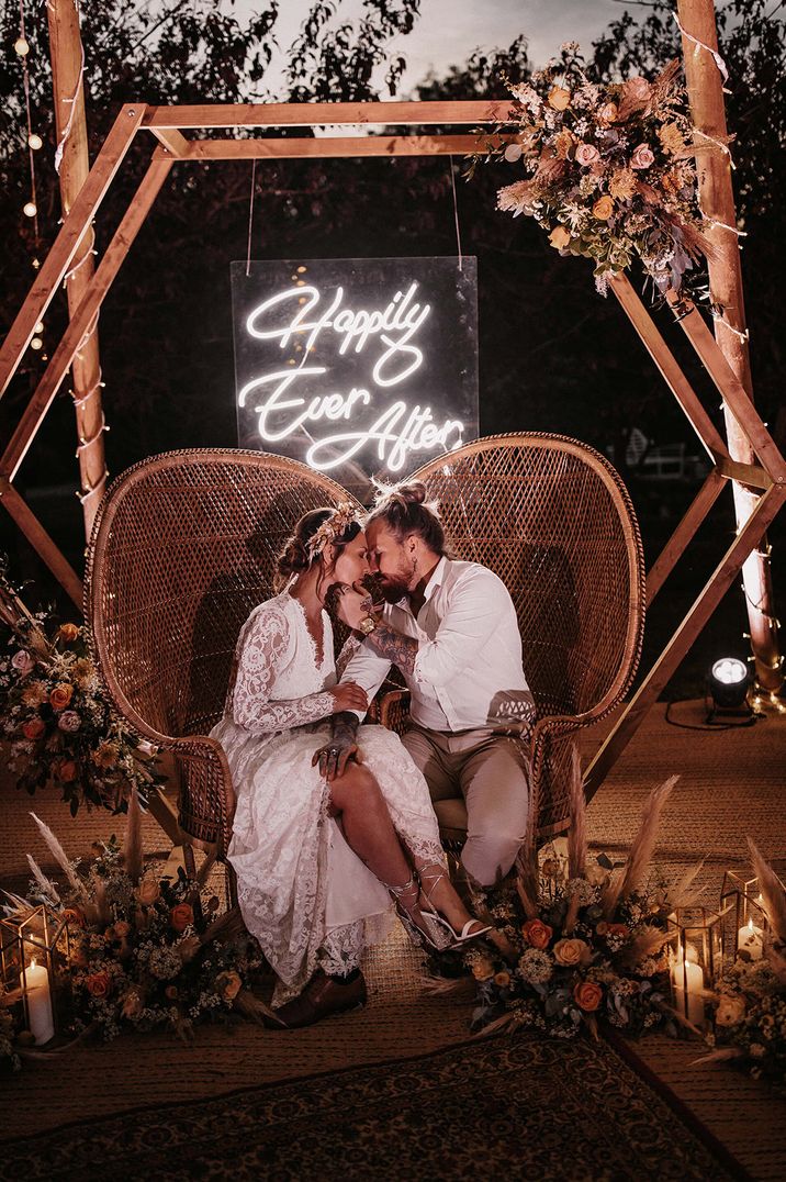 Bride and groom seated on peacock chairs underneath a white neon light 