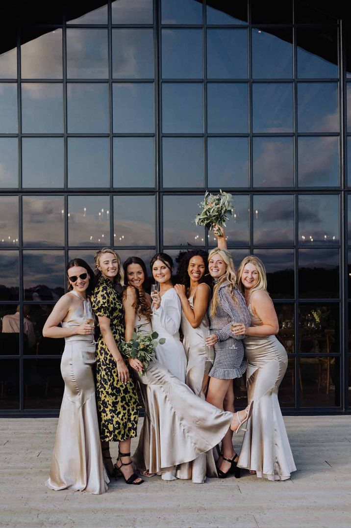 Bridal party in mismatched bridesmaid dresses standing outside floor-to-ceiling window of The Barn at Botley Hill wedding venue 