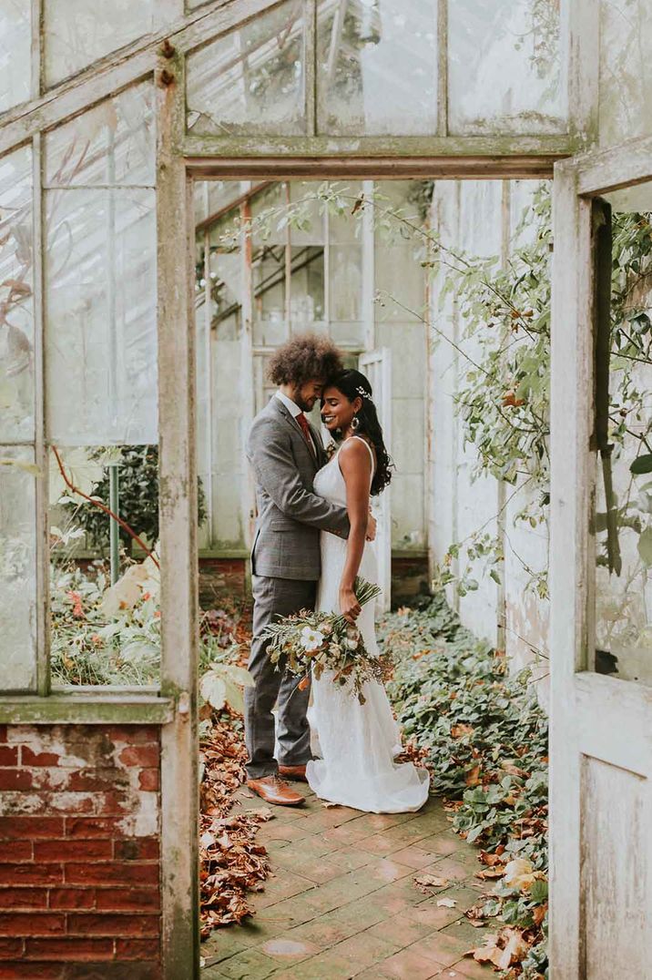 Bride in sleeveless lace wedding dress holding wildflower bouquet being embraced by groom in three piece grey suit in the glasshouse of Hooton Pagnel Hall wedding venue