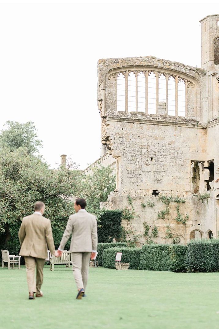 Grooms in light grey and tan grooms suits walking hand in hand on the grounds of Sudeley Castle wedding venue Gloucestershire 