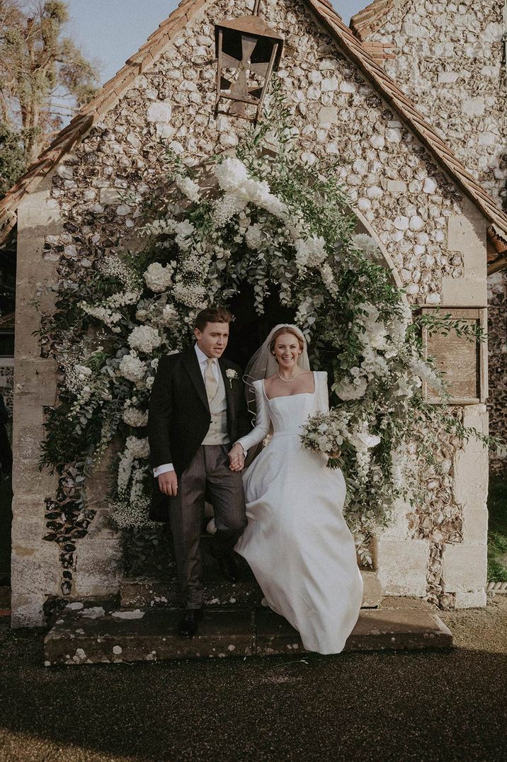 Bride in long sleeve wedding dress and groom under large white mixed floral arch at traditional wedding