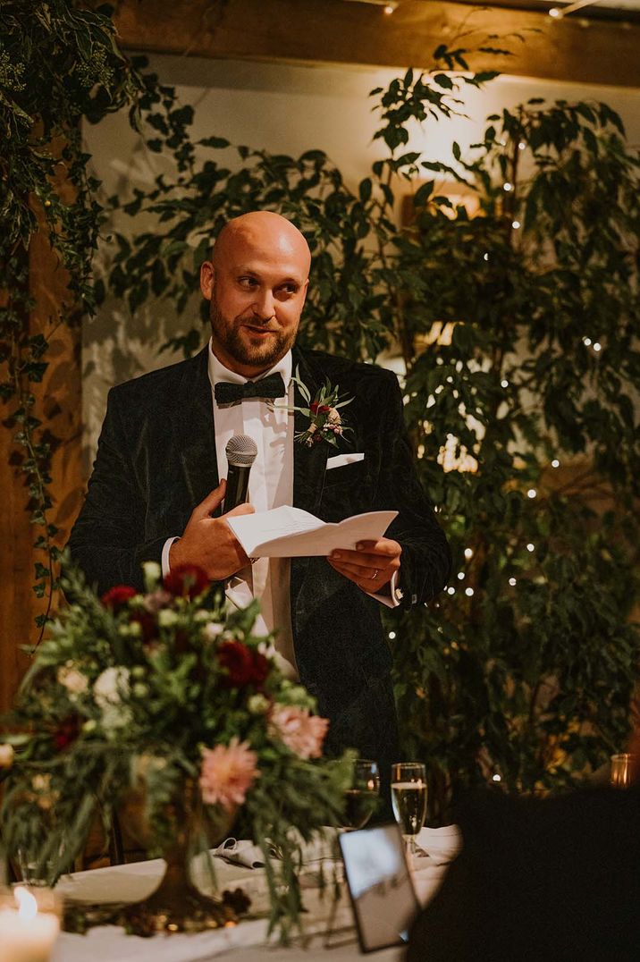 Groom in a velvet jacket and bow tie giving his groom speech at rustic barn reception