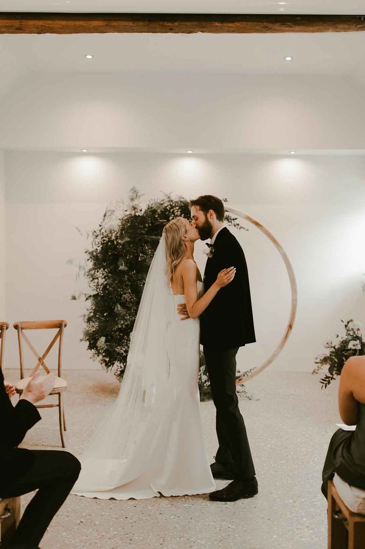 Bride and groom kissing at the alter at Southend Barns wedding venue with circular wooden arch with dried flower and foliage attached behind them 