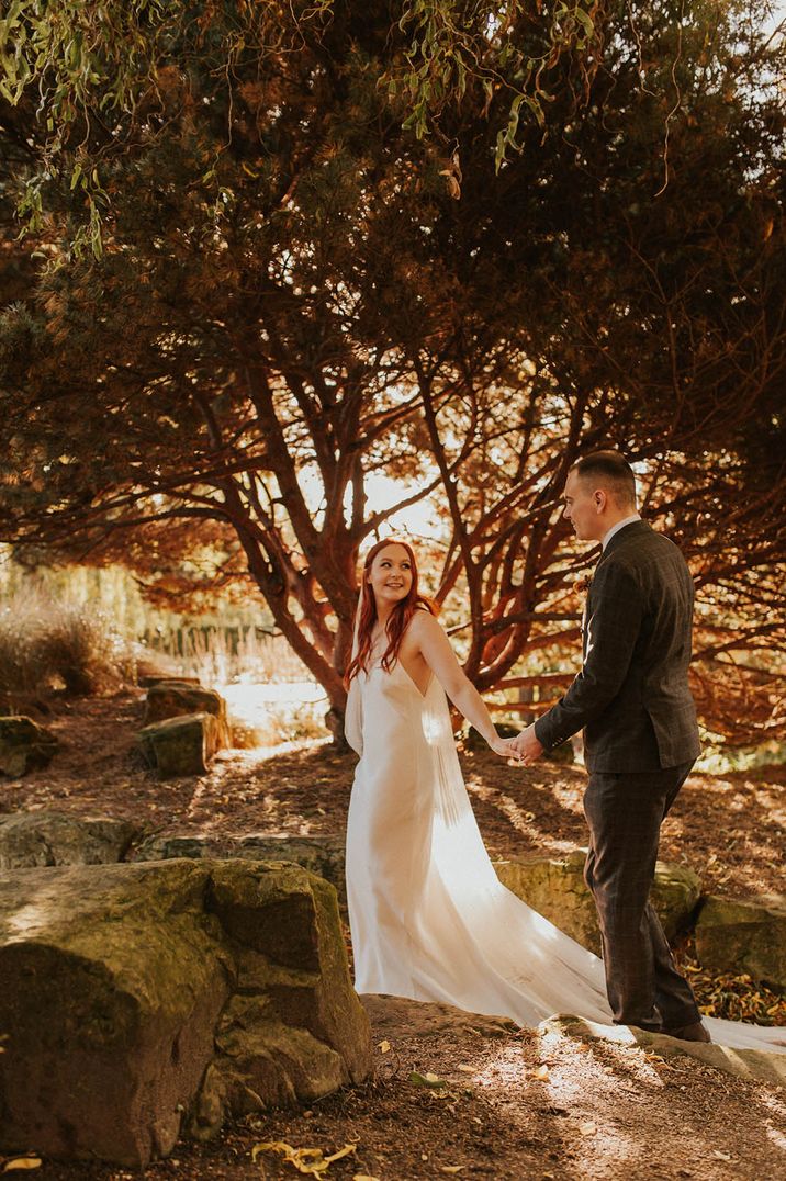 Bride at autumnal wedding in simple satin slip dress for wedding walking along with the groom 