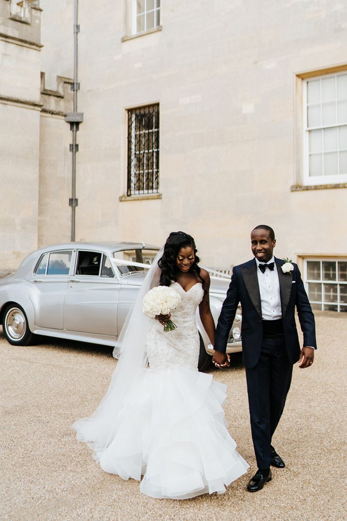 Bride in strapless wedding dress and groom holding hands with a vintage wedding car in the background