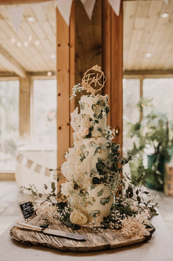 Extravagant three tier white wedding cake with gold flakes and lots of greenery and foliage with personalised cake topper