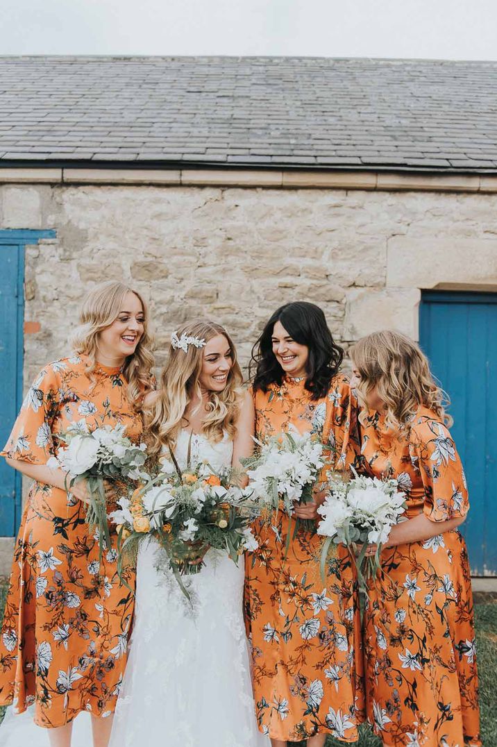 Bride in strapless lace wedding dress standing with bridesmaids in short sleeve midi orange floral patterned bridesmaid dresses