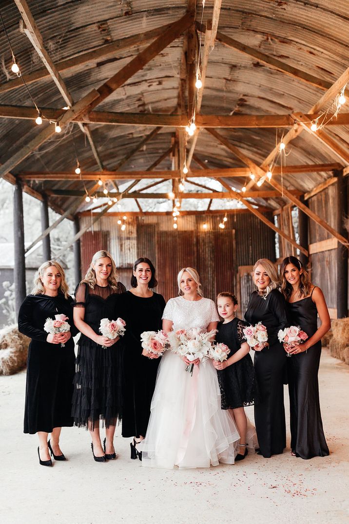 Bride standing with bridesmaids in black bridesmaid dresses in rustic barn winter wedding venue 