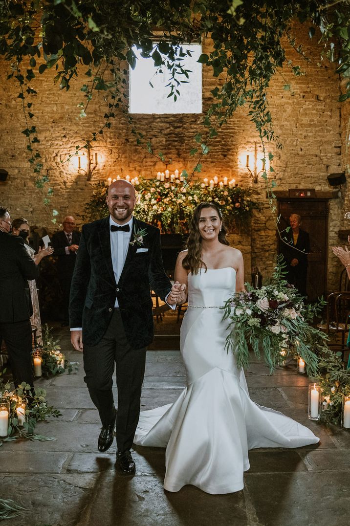 Bride in a strapless mermaid wedding dress descending down the rustic barn aisle decorated with green foliage with her husband in a black tuxedo 