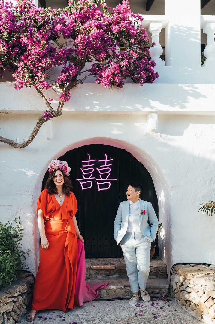 Newlyweds stand in front of pink LED light with Chinese symbols as they wear red gown and a pale blue suit 