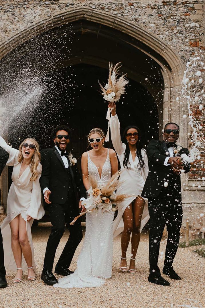 Champagne spray moment at a black-tie wedding with groomsmen in tuxedo, bridesmaids in satin dresses and bride in an appliqué wedding dress