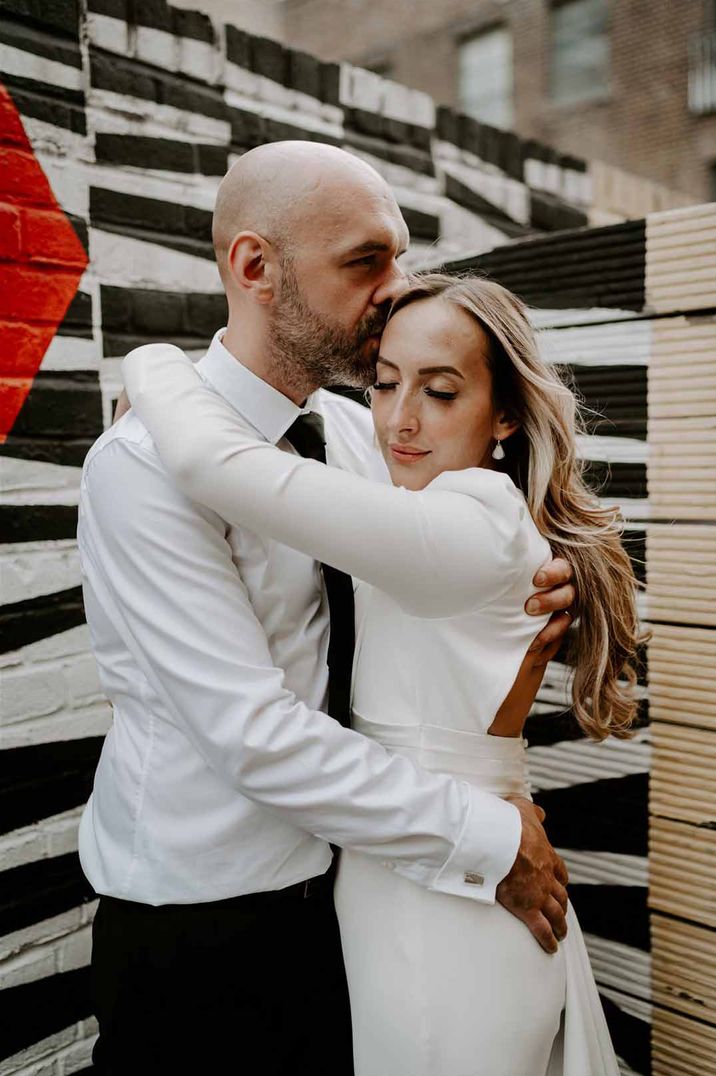 Bride in long sleeve wedding dress with open back detail embracing groom in white shirt and black tie at Shoreditch Studios - one of the wedding venues London 