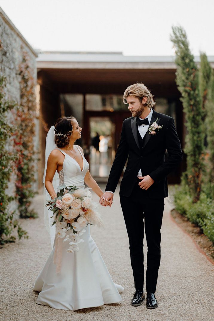 Bride carrying large oversized neutral wedding bouquet walking with groom in black tuxedo 