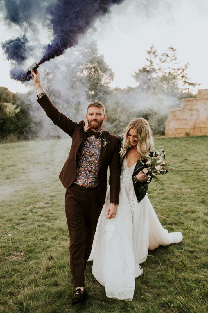 Bride in a princess wedding dress and leather jacket laughing with her groom in a floral shirt an brown blazer holding a smoke bomb