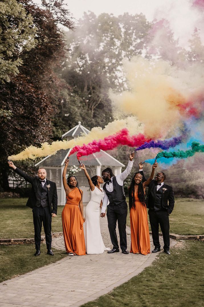 Groomsmen and groom wearing black tie with the bridesmaids in burnt orange satin dresses and bride in fitted off the shoulder wedding dress waving bright coloured smoke bombs