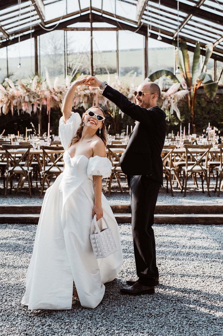 Bride wearing pearl handbag with white sunglasses being spun around by the groom in black tie for glasshouse wedding 