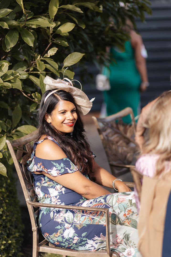 Wedding guest sitting down on a chair wearing a floral dress and wedding nude toned fascinator