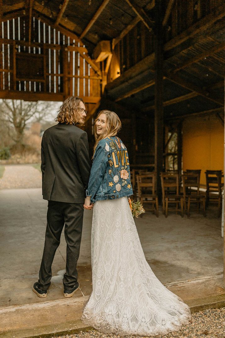 Bride wearing embroidered denim jacket with groom in black suit at barn wedding 