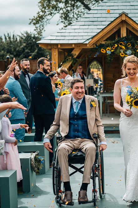 A bride and groom exit their wedding ceremony. The groom wears a light brown suit with blue checked waistcoat and is in a wheelchair. The bride wears a long white dress and carries a sunflower bouquet. 