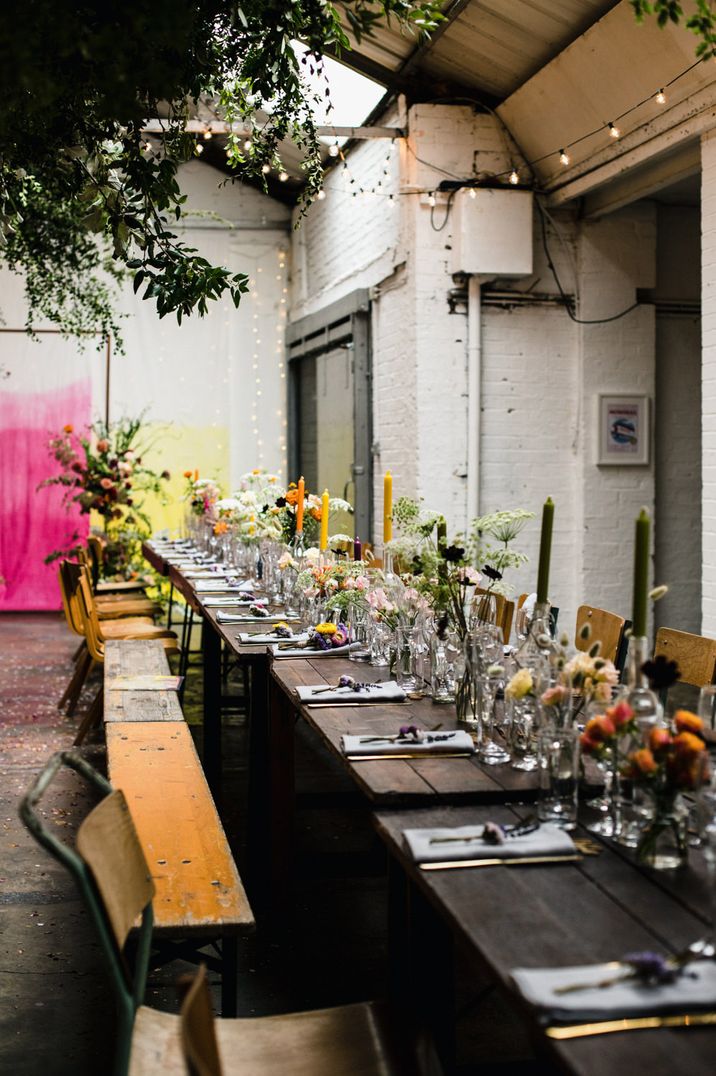 Industrial wedding venue with banquet tables littered with colourful wild flowers in vases and taper candles 