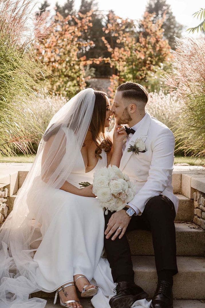 Bride in V neck wedding dress and veil holding white rose bouquet and groom in white double breasted tuxedo suit