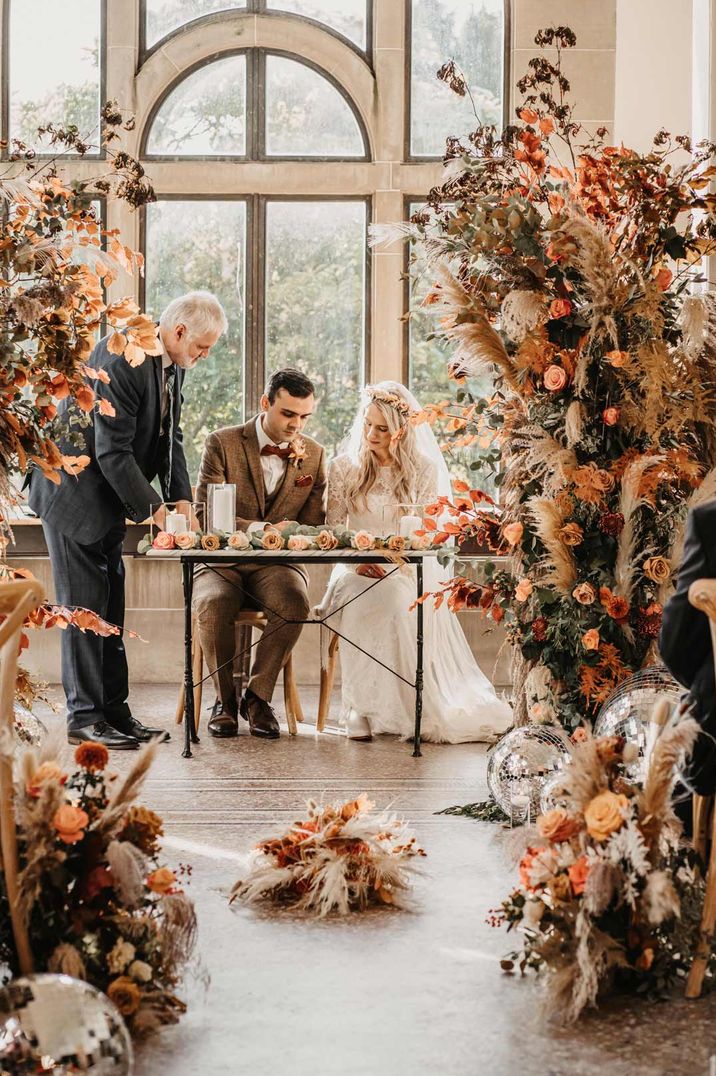 Bride in lace dress and flower crown and groom in orange themed wedding singing the register