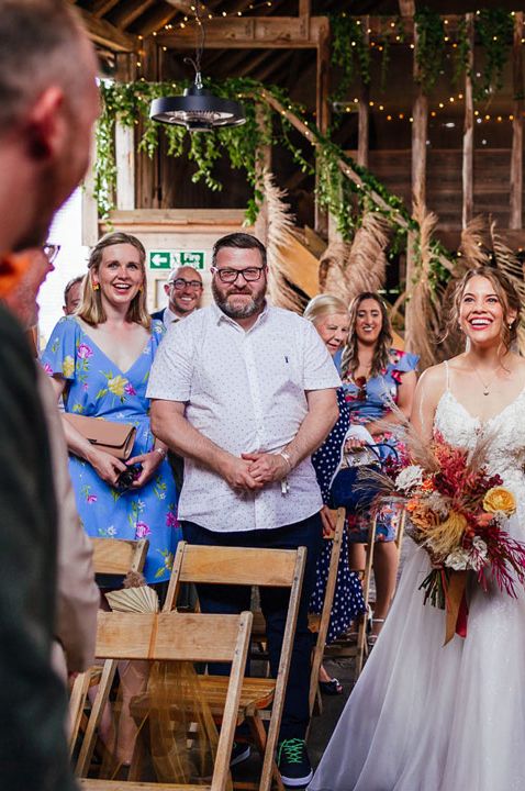 Bride in floral lace strappy wedding dress being walked down the aisle with vibrant floral bouquet