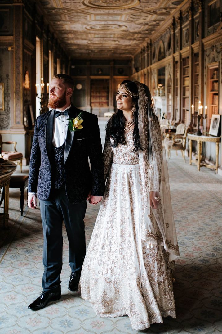 Bride wearing gold headdress and earrings with ivory and gold embroidered veil and dress with groom in black tie 