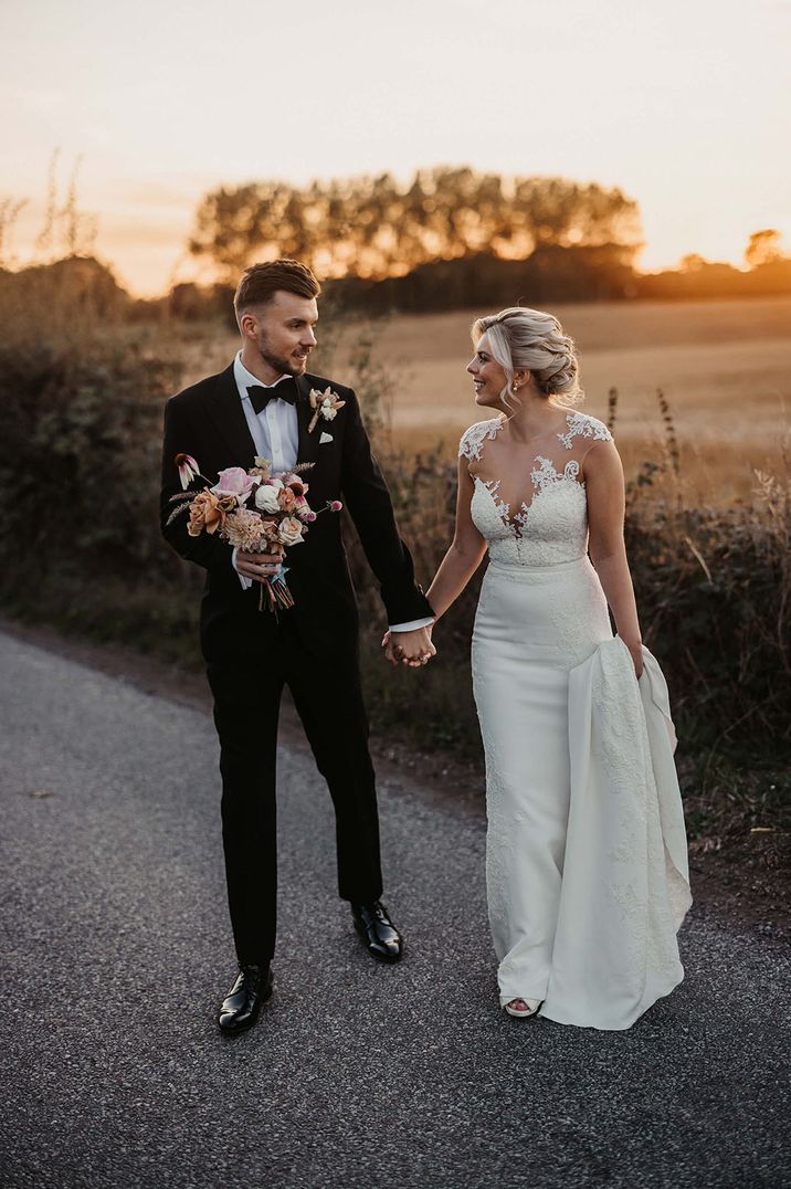 The groom in a black tuxedo stands with the bride during golden hour wearing a Pronovias illusion lace fitted wedding dress 