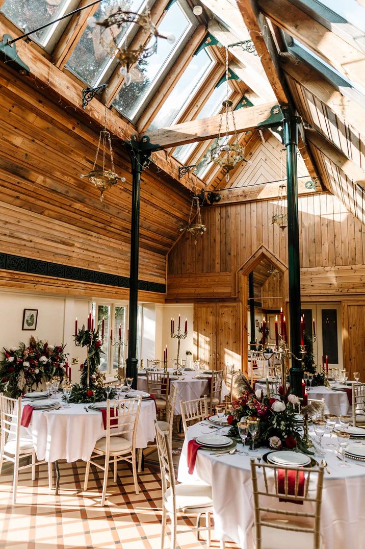 Reception room of Dunwood Hall Estate castle wedding venue with high ceilings, sky lights, black pillars, hanging lantern lighting, white tablecloths, red tapered candles and Christmas themed wedding floral centrepieces 