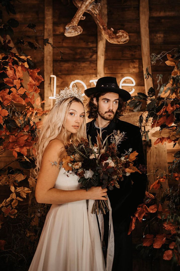 Halloween wedding with groom wearing hat and bride wearing dried flower crown standing between autumn flower columns and bridal bouquet