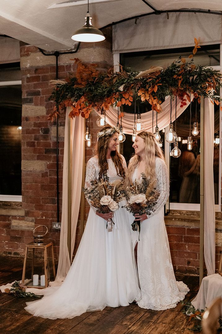 Brides in boho lace wedding dresses standing beneath their autumnal wedding decor with seasonal flowers and leaves 