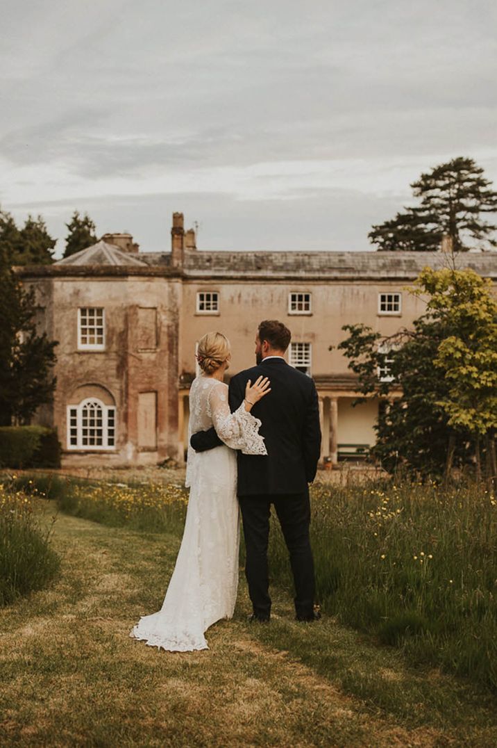 Bride in lace sleeve wedding dress and groom in dark suit embracing on the grounds of Pennard House, one of the best UK country house wedding venues