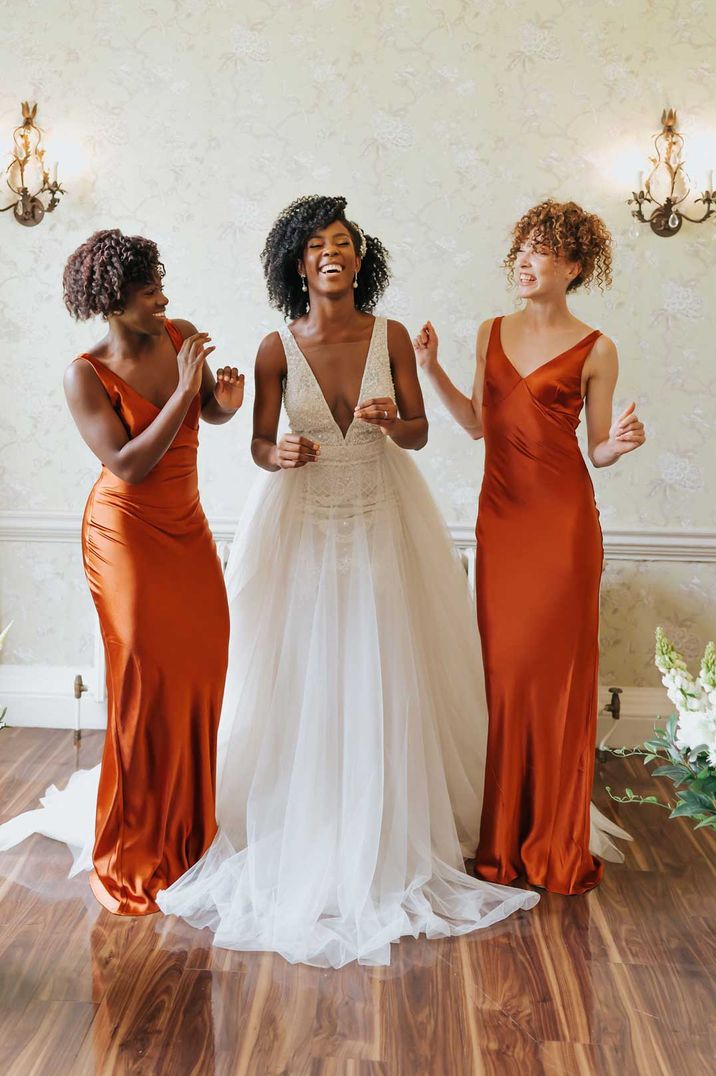 Bride in plunging neckline sleeveless wedding dress standing with two bridesmaids in satin orange bridesmaid dresses