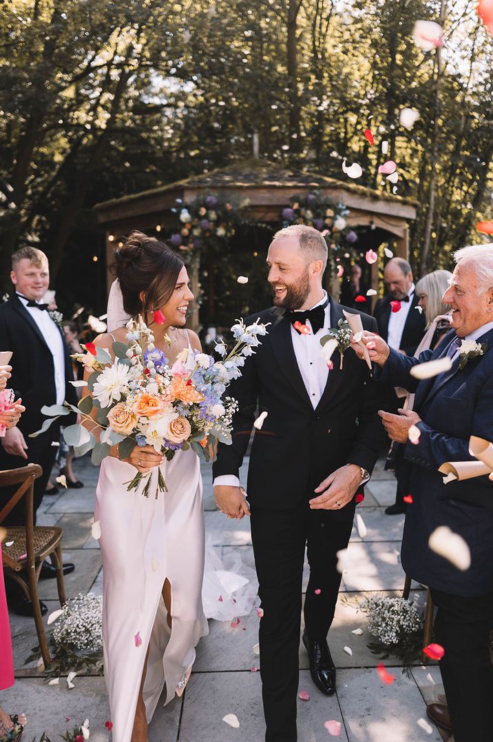 Groom in black tuxedo walking out with the bride in a slip wedding dress at their outdoor ceremony at The Venue Barkisland 