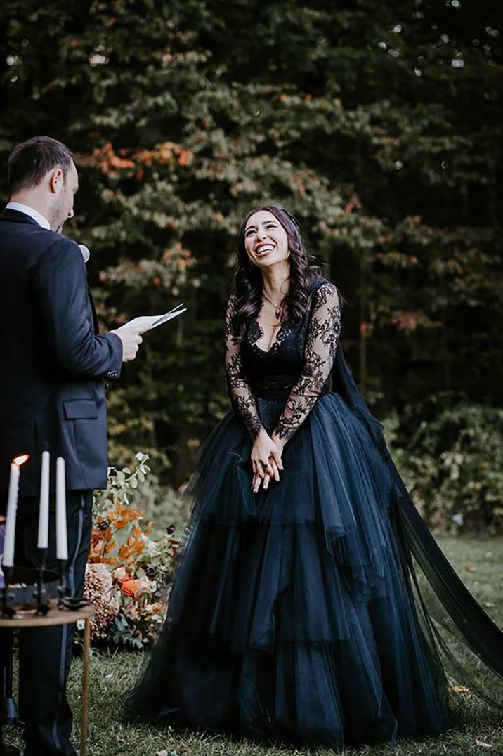 Bride wearing a black tulle layered wedding dress smiles at the groom during their wedding vows at outdoor wedding ceremony in New York for gothic wedding 