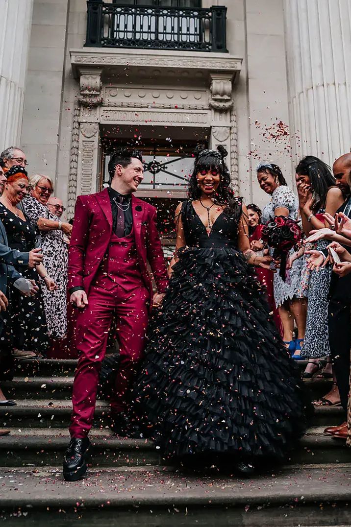 Bride in grand black ruffled wedding dress and groom in maroon grooms suit walking down the steps of a town hall wedding venue