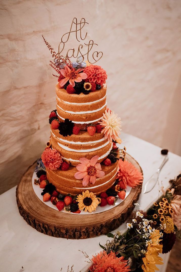 Wire wedding cake topper sits on a three tier naked wedding cake with colourful flowers and summer berries