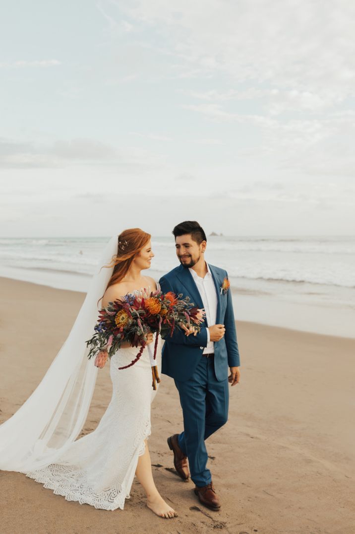 Bride and groom walking along the beach with orange wedding bouquet