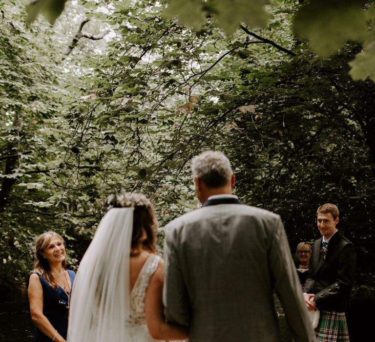 Father of the bride walks the bride down the aisle towards her groom awaiting her in traditional Scottish dress