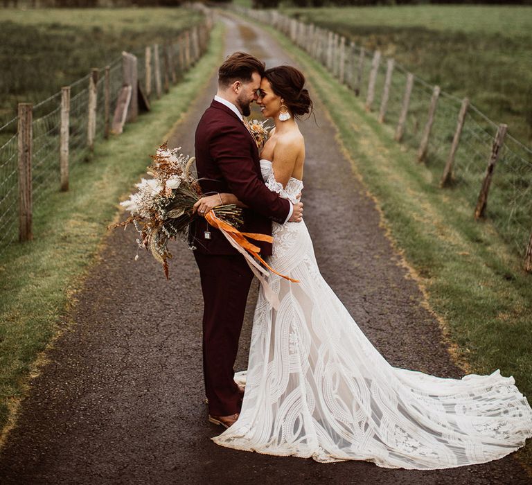 Smiling boho bride holding pampas grass bouquet embraces groom in burgundy suit in the Lake District