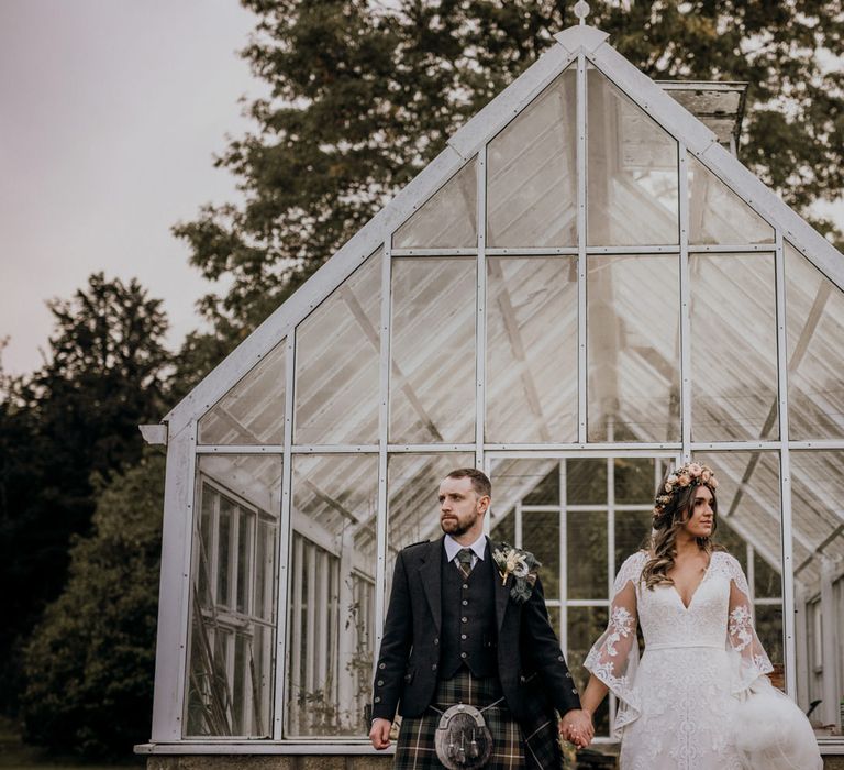 Boho bride in rose flower crown and groom in tartan kilt hold hands in front of greenhouse at highland wedding in Glencoe