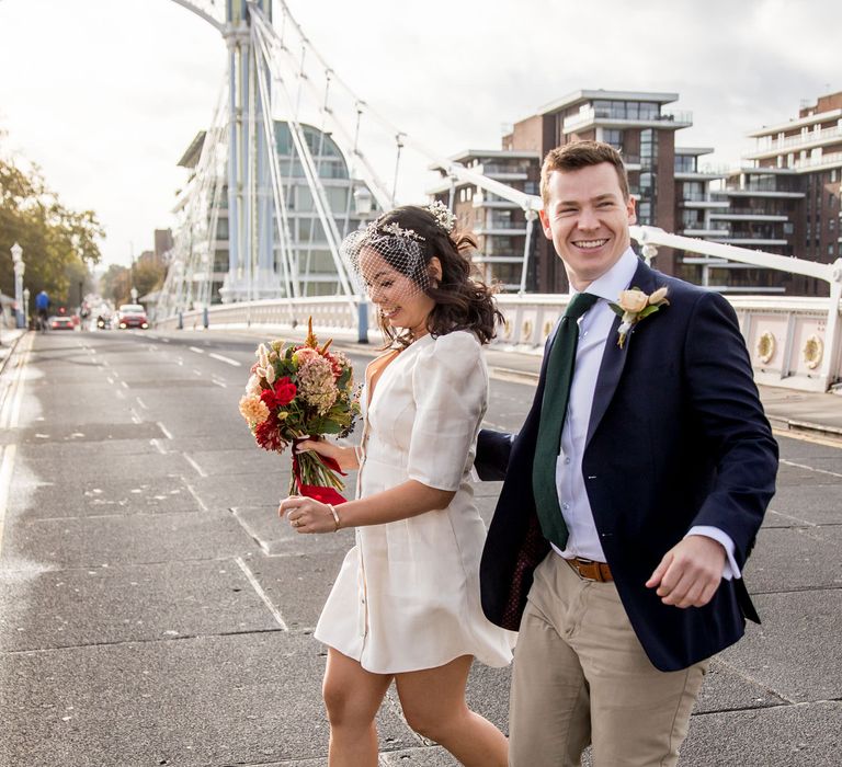 Bride in short registry wedding dress and groom in chinos and blazer walking across Albert Bridge in London 